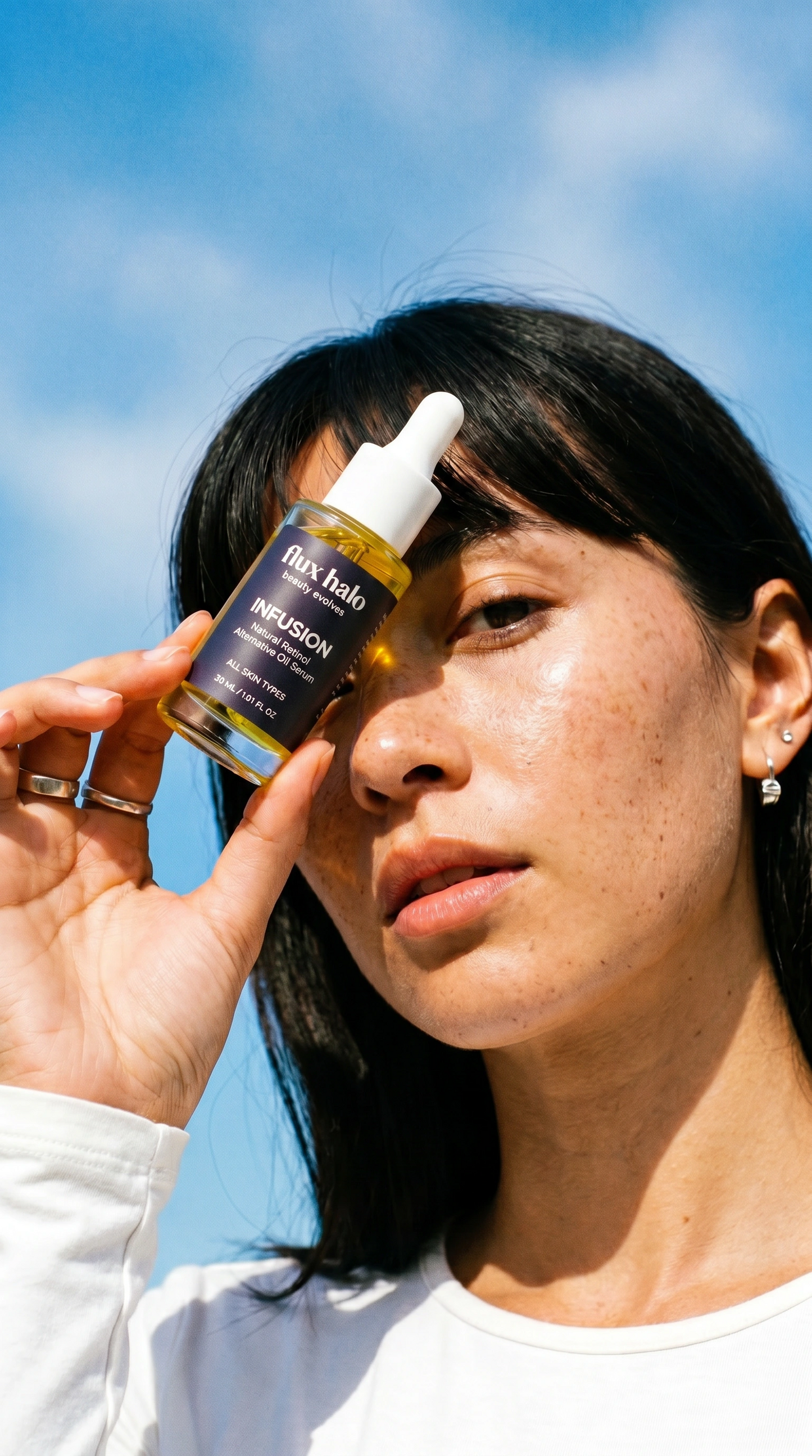 A woman outdoors holding a small dropper bottle of hair oil near her face against a bright blue sky. The bottle label reads 'flux hair' and 'MIPSION'.